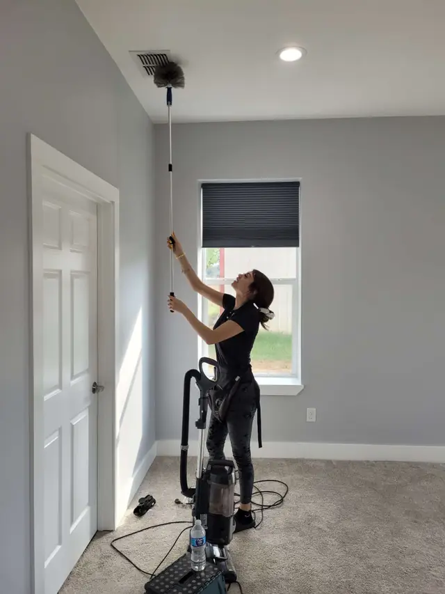 A technician on an extension pole dusting a ceiling vent in an emptied room, a vacuum at her feet
