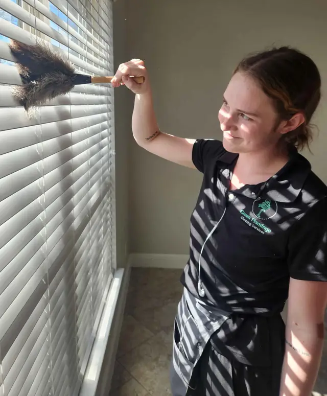 A technician in the black Green Flamingo polo dusting the slats of a set of window blinds