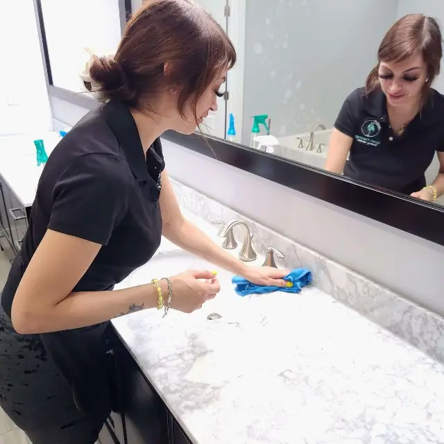 A Green Flamingo technician wiping down a marble bathroom counter, the mirror behind her